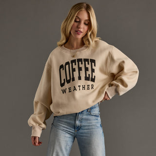 A woman wearing a beige crewneck sweatshirt with 'COFFEE WEATHER' printed in black block letters on the front, paired with light blue jeans, against a solid dark gray studio background.