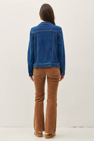A woman viewed from behind wearing a blue denim jacket and brown corduroy flare pants against a plain white studio background.