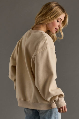A woman wearing an oversized beige crewneck sweatshirt, shown from a side-back angle against a solid gray studio background.