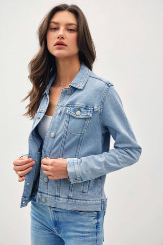 A woman wearing a light blue denim jacket over a white top and matching blue jeans, photographed against a plain light gray studio background.