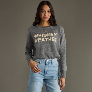 Woman wearing a gray sweater with 'WHISKEY WEATHER' text and blue jeans against a gray background