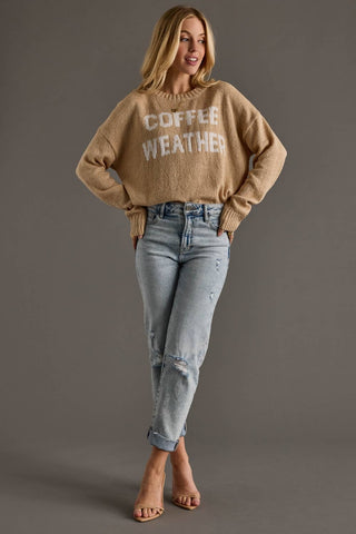 A woman modeling a beige crewneck sweater with 'COFFEE WEATHER' written in white varsity-style letters across the chest, paired with light-wash blue jeans and clear high-heel sandals, against a solid gray studio background.