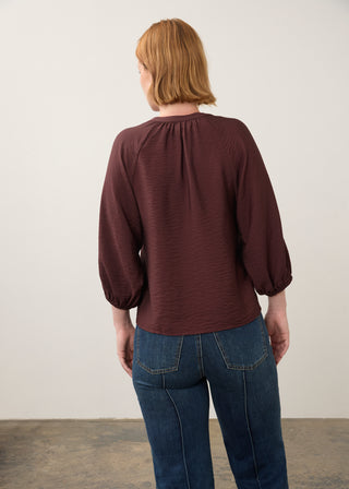 A woman viewed from the back wearing a dark burgundy long-sleeve blouse with a gathered neckline and blue jeans, standing against a plain off-white wall.