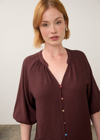 A woman with short red hair wearing a dark brown crinkled blouse with a split neckline and decorative buttons, photographed against a plain light background.