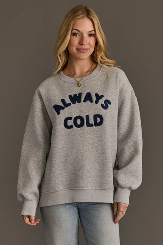 Woman wearing a gray sweatshirt with 'ALWAYS COLD' text on a plain background
