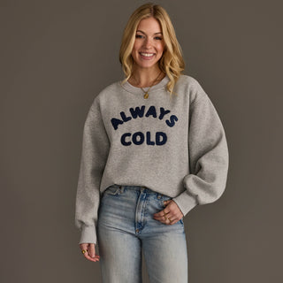 A woman wearing a light gray crewneck sweatshirt with 'ALWAYS COLD' printed in navy blue across the chest, paired with light-wash blue jeans, against a solid dark gray studio background.