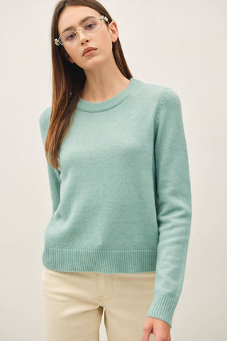 A woman wearing a light mint green crewneck knit sweater and cream-colored pants, photographed against a plain light gray studio background.