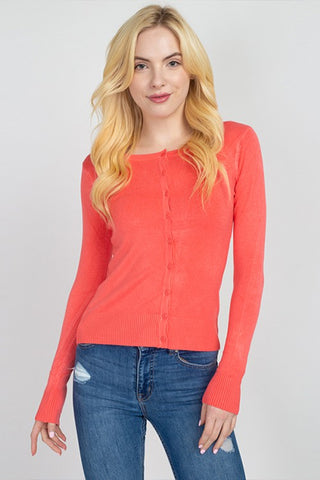 A blonde woman wearing a coral pink long-sleeve knit cardigan with front buttons and blue jeans, standing against a light gray studio background.