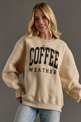 A woman wearing a beige crewneck sweatshirt with 'COFFEE WEATHER' printed in black varsity-style letters on the front, paired with light blue jeans, against a solid dark gray studio background.