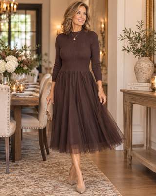 A woman models a long-sleeve ribbed knit midi dress in a warm brown color, featuring a flared tulle skirt. She is standing in a beautifully decorated dining room with a table, chairs, and a console table in the background.