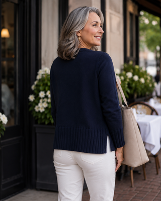 A woman with gray hair is shown from behind wearing a navy blue knit sweater and white pants, carrying a beige tote bag in front of a cafe storefront.