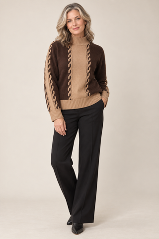 A woman modeling a brown and beige cable-knit mock-neck sweater with black wide-leg trousers, standing against a solid beige studio background.