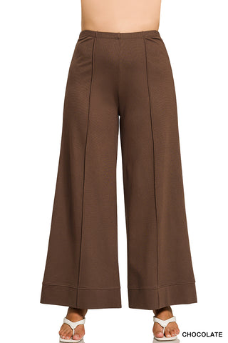 A model is wearing high-waisted, wide-leg cropped pants in a chocolate brown color, paired with white heeled sandals, shown against a plain white background.