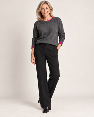 A woman modeling a dark gray crewneck sweater with bright pink trim at the collar and cuffs, paired with black wide-leg trousers, against a light beige studio background.