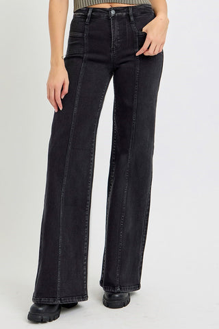A person wearing black wide-leg jeans with front seam detailing, shown from the waist down against a light gray studio background.
