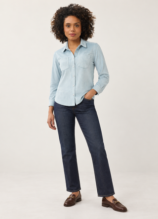 A woman modeling a light blue long-sleeve button-up shirt and dark blue straight-leg jeans with brown leather loafers, standing against a plain light gray studio background.