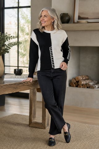 A woman stands in a modern living room wearing a color-block knit sweater with black and off-white panels, black wide-leg jeans, and black loafers with gold hardware.
