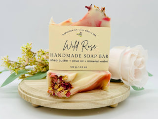 Handmade soap bar with 'Wild Rose' branding on a wooden stand with flowers in the background.