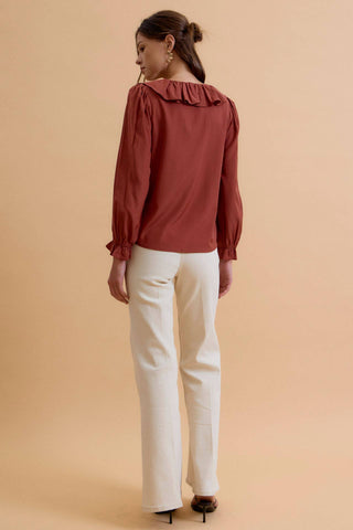 A woman shown from the back wearing a rust-colored long-sleeve blouse with a ruffled collar and white trousers, standing against a solid beige background.