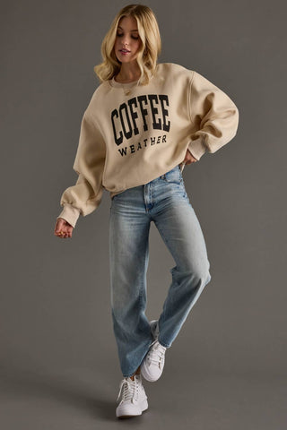 A woman modeling a beige crewneck sweatshirt with 'COFFEE WEATHER' printed in black on the front, paired with light-wash blue jeans and white sneakers, against a solid gray studio background.