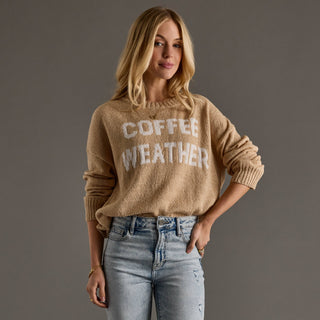 A woman wearing a beige fuzzy knit sweater with 'COFFEE WEATHER' written in white across the chest, paired with light-wash blue jeans, against a solid dark gray studio background.