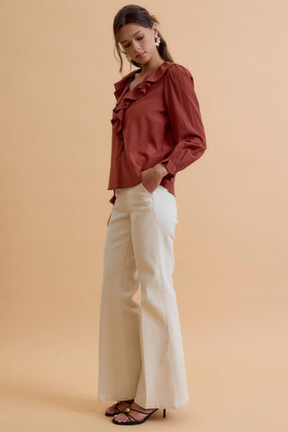 A woman modeling a rust-brown ruffled blouse with long sleeves, paired with cream wide-leg trousers and black strappy heels, set against a solid warm beige studio background.