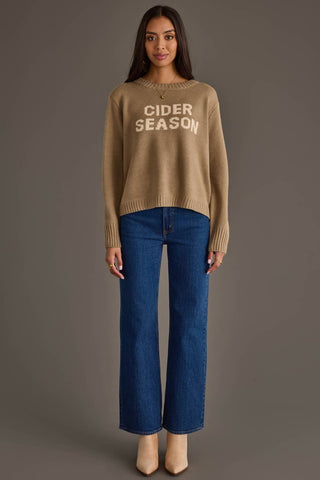 A woman stands against a plain taupe studio background wearing a beige crewneck sweater with 'CIDER SEASON' printed in white across the chest, paired with dark blue wide-leg jeans and beige heels.