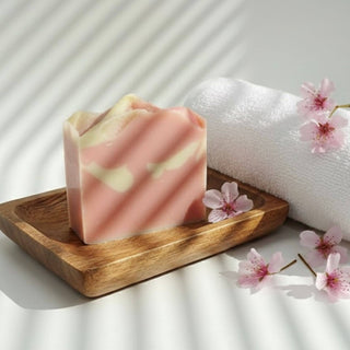 Pink soap bar on a wooden tray with cherry blossoms and a towel in the background
