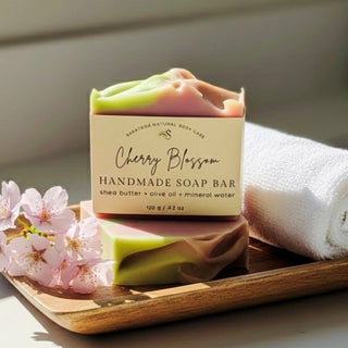 Handmade soap bar with cherry blossom design on a wooden tray with flowers and towel.