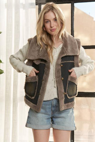 A woman modeling a brown and black shearling-style vest with large front pockets, worn over a cream sweater and denim shorts in a softly lit indoor setting.