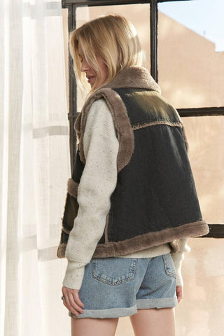A woman wearing a dark gray denim vest with brown shearling-style trim, layered over a cream knit sweater and paired with light blue denim shorts, standing near a window.