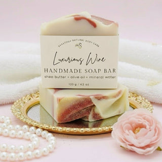Handmade soap bar with packaging on a gold plate, surrounded by pearls and a pink flower on a soft pink background.