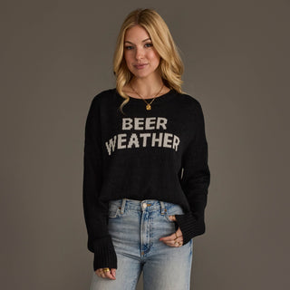A woman modeling a black knit sweater with 'BEER WEATHER' written across the chest in white varsity-style letters, paired with light blue jeans against a solid taupe background.