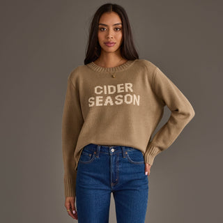 A woman modeling a tan knit sweater with 'CIDER SEASON' text across the chest, paired with blue jeans, against a solid gray studio background.