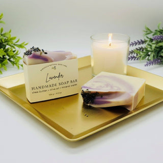 Handmade lavender soap bar on a gold tray with a lit candle and greenery in the background.