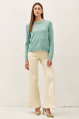A woman modeling a mint green crewneck knit sweater and cream-colored wide-leg trousers against a plain off-white studio background.