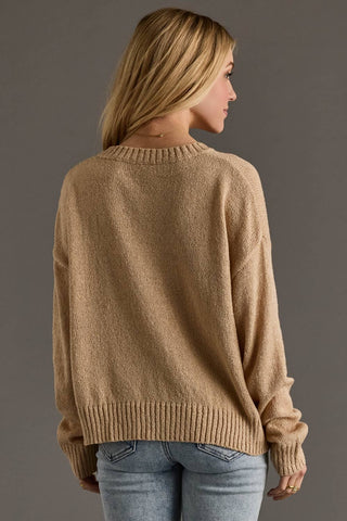 A woman shown from the back wearing a loose, light brown knit sweater and light blue jeans against a solid dark gray studio background.