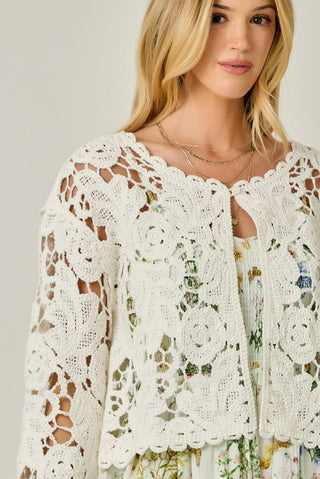 Woman wearing a white lace cardigan over a floral dress against a neutral background