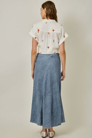 Woman wearing a white blouse with colorful patterns and a long denim skirt on a plain background