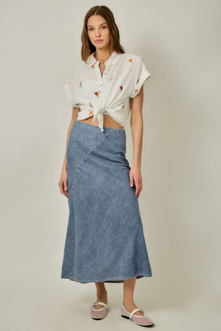Woman wearing a white blouse with floral patterns and a blue denim skirt on a plain background